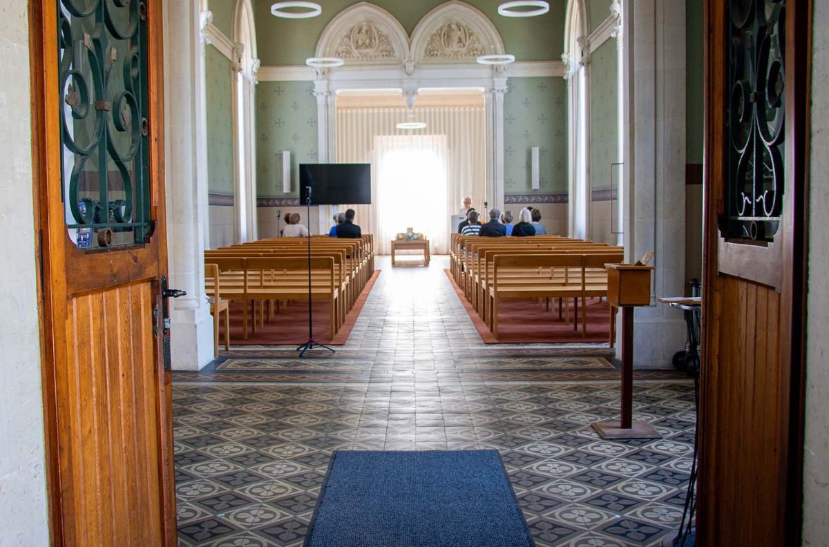 Funeral ceremony in a chapel viewed through open doors, with screen and live streaming setup